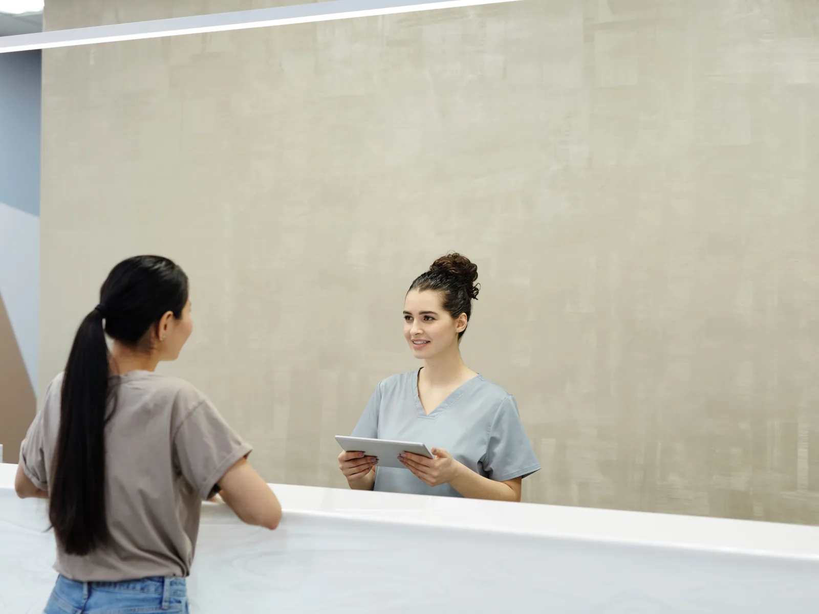 A receptionist uses a tablet while assisting a patient at a clinic reception desk.