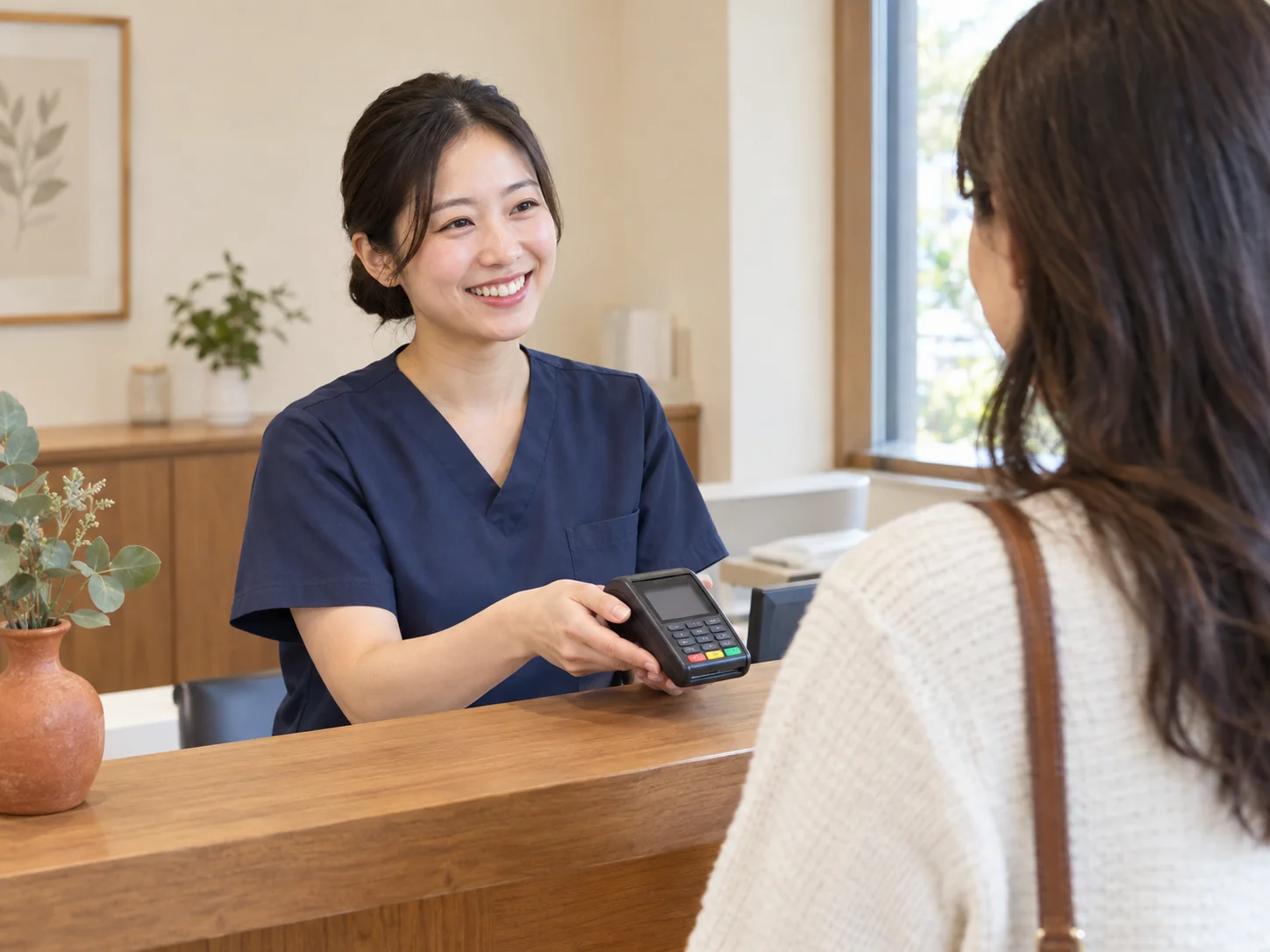 A smiling receptionist at a warm wood dental reception counter holds a payment terminal for a patient.