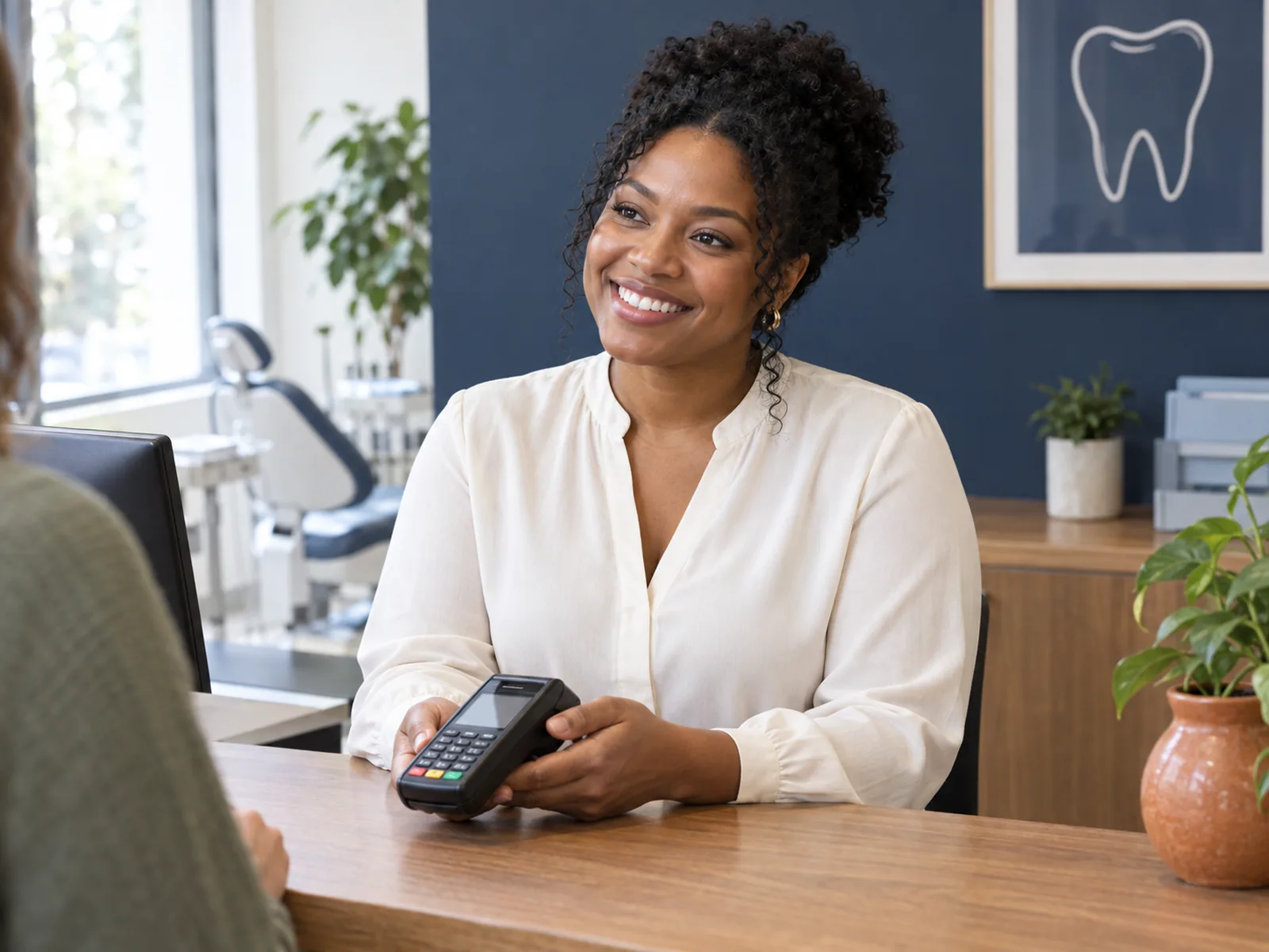 A smiling receptionist at a warm wood dental reception counter holds a payment terminal for a patient.